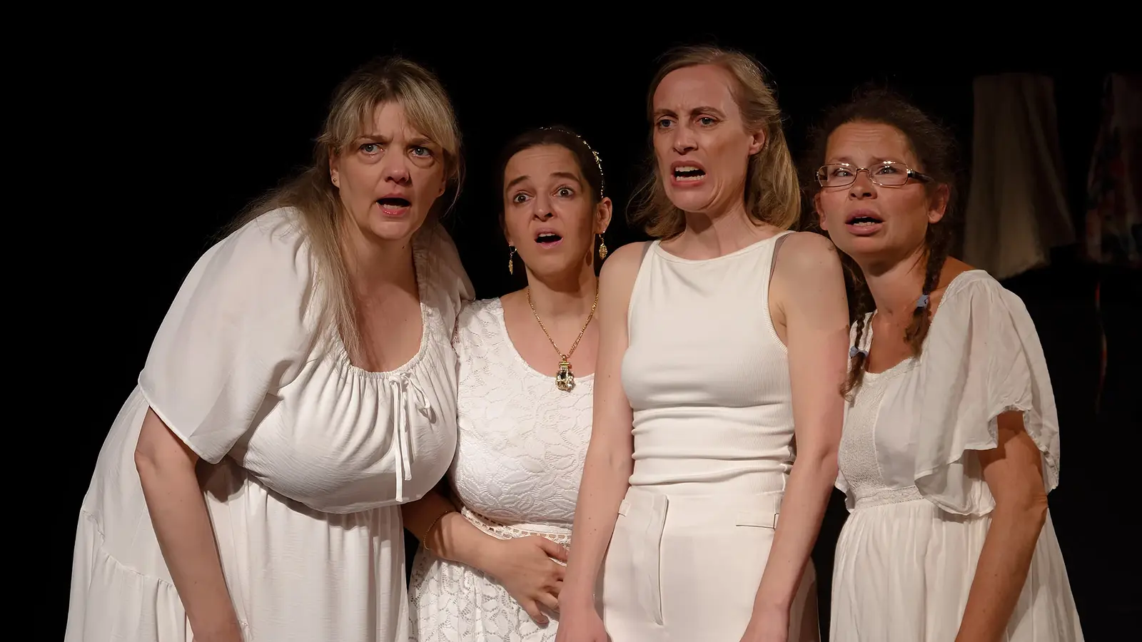 Bühnenfoto aus Lysistrata mit vier Darstellenden in weißen Kostümen vor schwarzem Bühnenhintergrund, die dicht beieinanderstehen und nach vorne blicken, eine Person mit Brille und drei weitere in unterschiedlichen weißen Kleidern