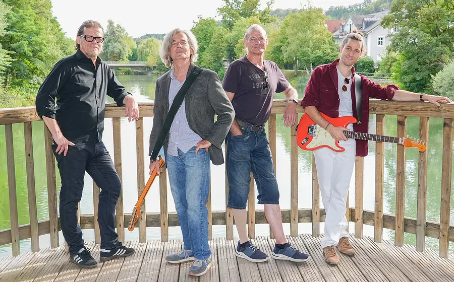 Bandfoto von Magic Buzz mit vier Musikern auf einer Holzplattform am Fluss. Zwei Personen halten E-Gitarren, im Hintergrund sind Bäume und Wohnhäuser zu sehen.