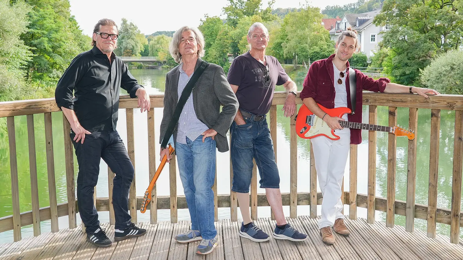 Bandfoto von Magic Buzz mit vier Musikern auf einer Holzplattform am Fluss. Zwei Personen halten E-Gitarren, im Hintergrund sind Bäume und Wohnhäuser zu sehen.
