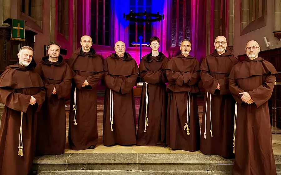 Acht Sänger in braunen Mönchskutten stehen in einer Kirche vor einem beleuchteten Kreuz und farbigem Bühnenlicht