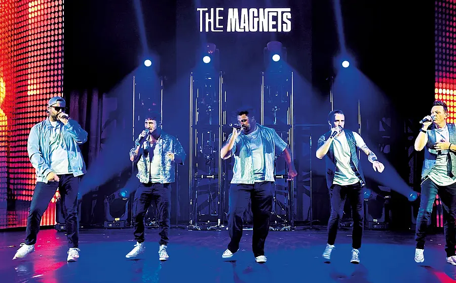 Bühnenfoto The Magnets mit fünf Sängern die mit Mikrofonen in einer Reihe auf beleuchteter Bühne auftreten vor LED Wänden und Schriftzug The Magnets