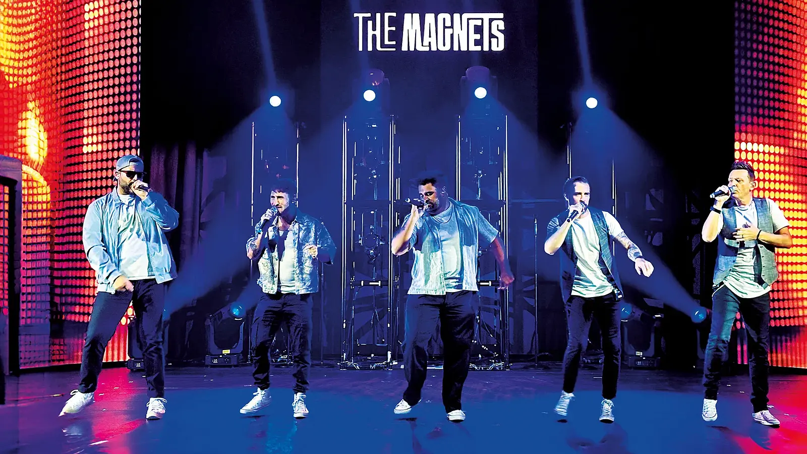 Bühnenfoto The Magnets mit fünf Sängern die mit Mikrofonen in einer Reihe auf beleuchteter Bühne auftreten vor LED Wänden und Schriftzug The Magnets