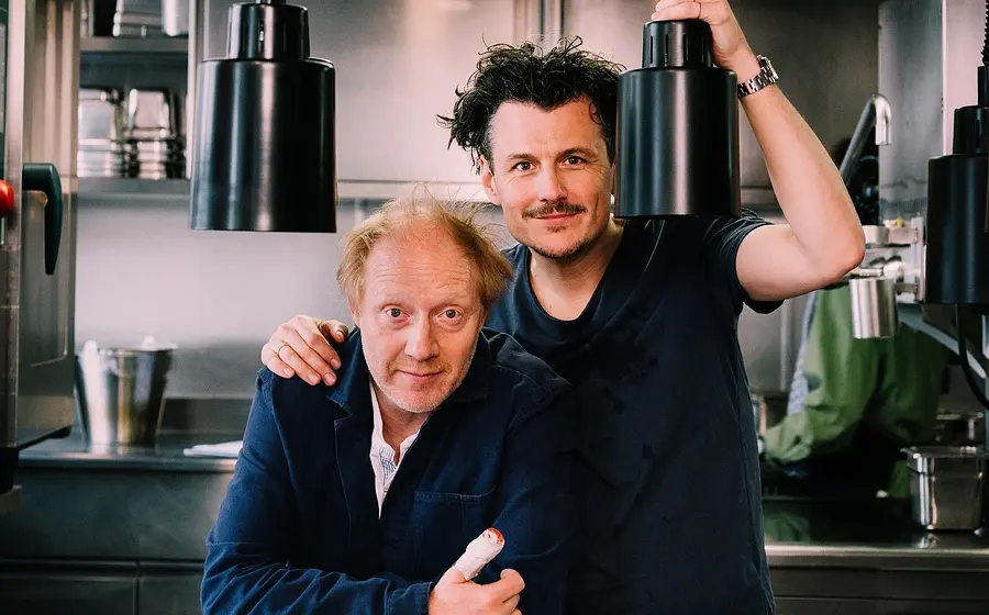 Manuel Rubey und Simon Schwarz in einer Küche mit schwarzen Hängelampen und moderner Einrichtung