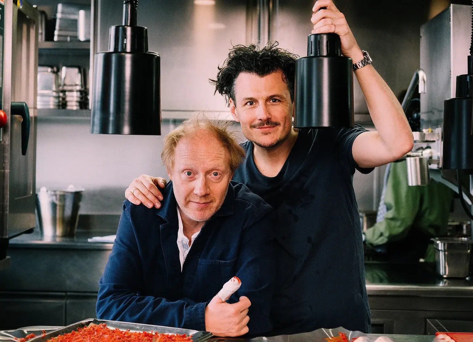 Manuel Rubey und Simon Schwarz in einer Küche mit schwarzen Hängelampen und moderner Einrichtung