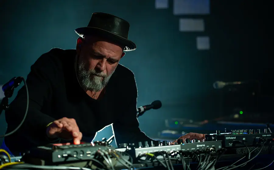 Martin Kälberer mit schwarzem Hut an einem Mischpult und elektronischem Equipment, bedient Regler und Geräte auf einer abgedunkelten Bühne mit blauem Licht und Kabeln im Vordergrund.