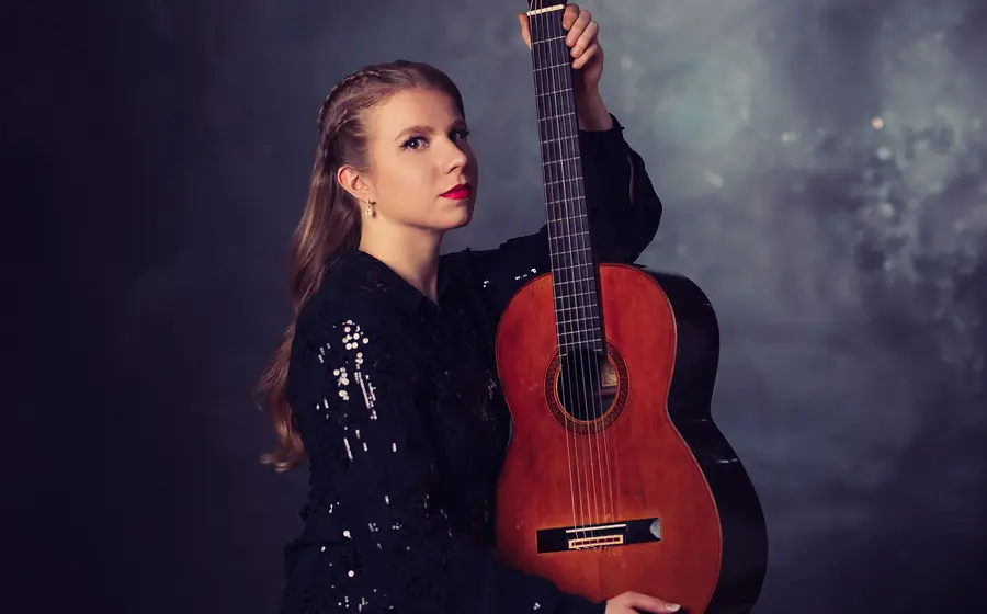 Eine Person in schwarzem, glitzerndem Oberteil mit klassischer Gitarre vor dunkelblauem Studiohintergrund, die das Instrument aufrecht hält und direkt zur Kamera blickt.