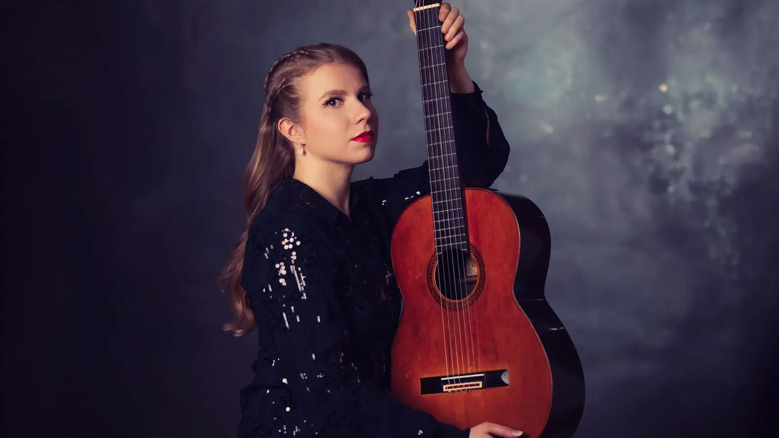 Eine Person in schwarzem, glitzerndem Oberteil mit klassischer Gitarre vor dunkelblauem Studiohintergrund, die das Instrument aufrecht hält und direkt zur Kamera blickt.
