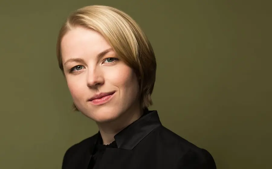 Porträtfoto mit Person in schwarzer Kleidung vor olivgrünem Hintergrund, in Nahaufnahme mit kurzem blondem Haar und direktem Blick zur Kamera