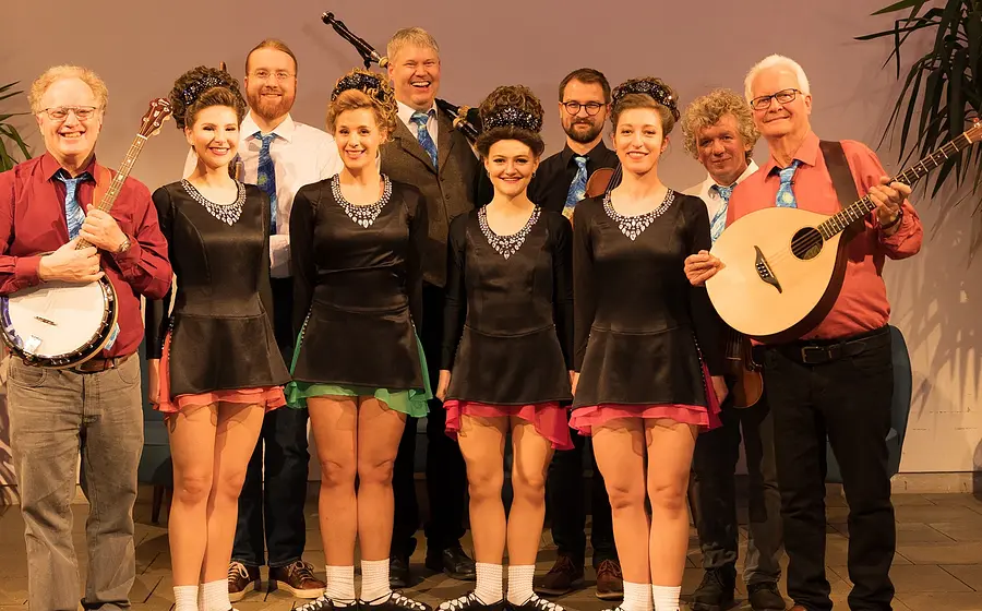 Musikensemble mit Instrumentalisten und Tänzern steht in Reihe auf Bühne, einige halten Banjo und Mandoline, mehrere tragen koordinierte Bühnenkostüme.