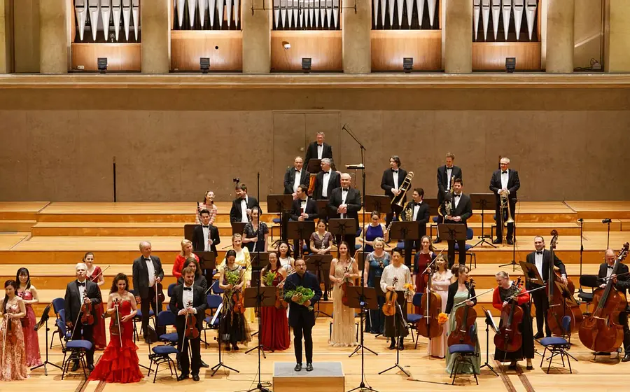 Großes Orchester auf einer Konzertbühne: Dirigent vorne mittig, Musiker mit Streich- und Blasinstrumenten sowie einegroße Orgel im Hintergrund.