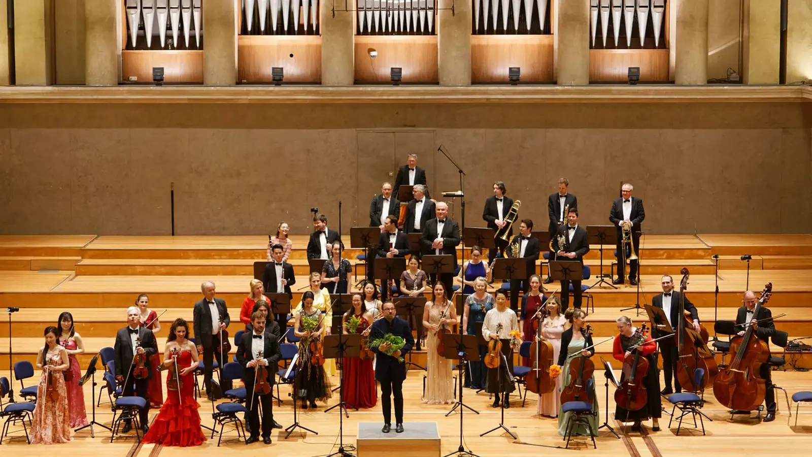 Großes Orchester auf einer Konzertbühne: Dirigent vorne mittig, Musiker mit Streich- und Blasinstrumenten sowie einegroße Orgel im Hintergrund.