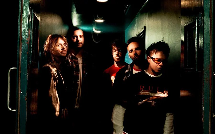 Bandfoto von fünf Musikern in einem dunklen, atmosphärisch beleuchteten Gang mit dramatischem Licht und Indie-Rock-Ästhetik.