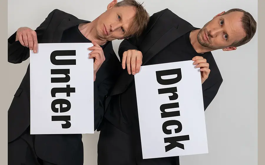 Foto von zwei Personen in schwarzen Outfits vor hellem Hintergrund, die weiße Tafeln mit den Aufschriften „Unter“ und „Druck“ halten und ihre Köpfe gegeneinander lehnen.