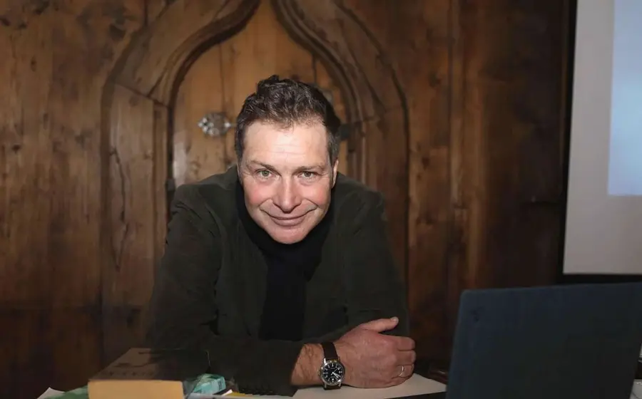 Oliver Pötsch in dunklem Sakko und schwarzem Rollkragenpullover, die auf einem Tisch vor einer geschnitzten Holzwand lehnt, neben Büchern und einem Laptop in einem holzvertäfelten Raum