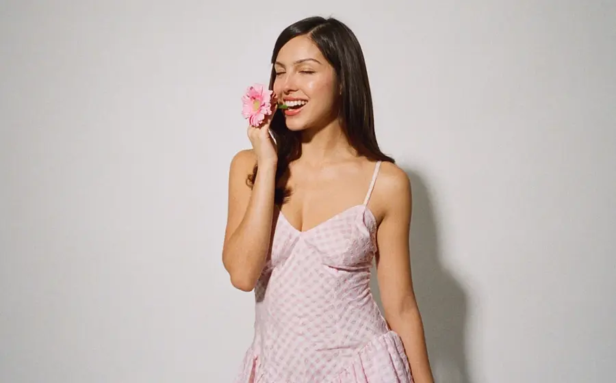 Olivia Rodrigo in rosafarbenem Kleid hält eine Blume an ihr Gesicht vor neutralem Hintergrund, offizielles Pressefoto der Sängerin.