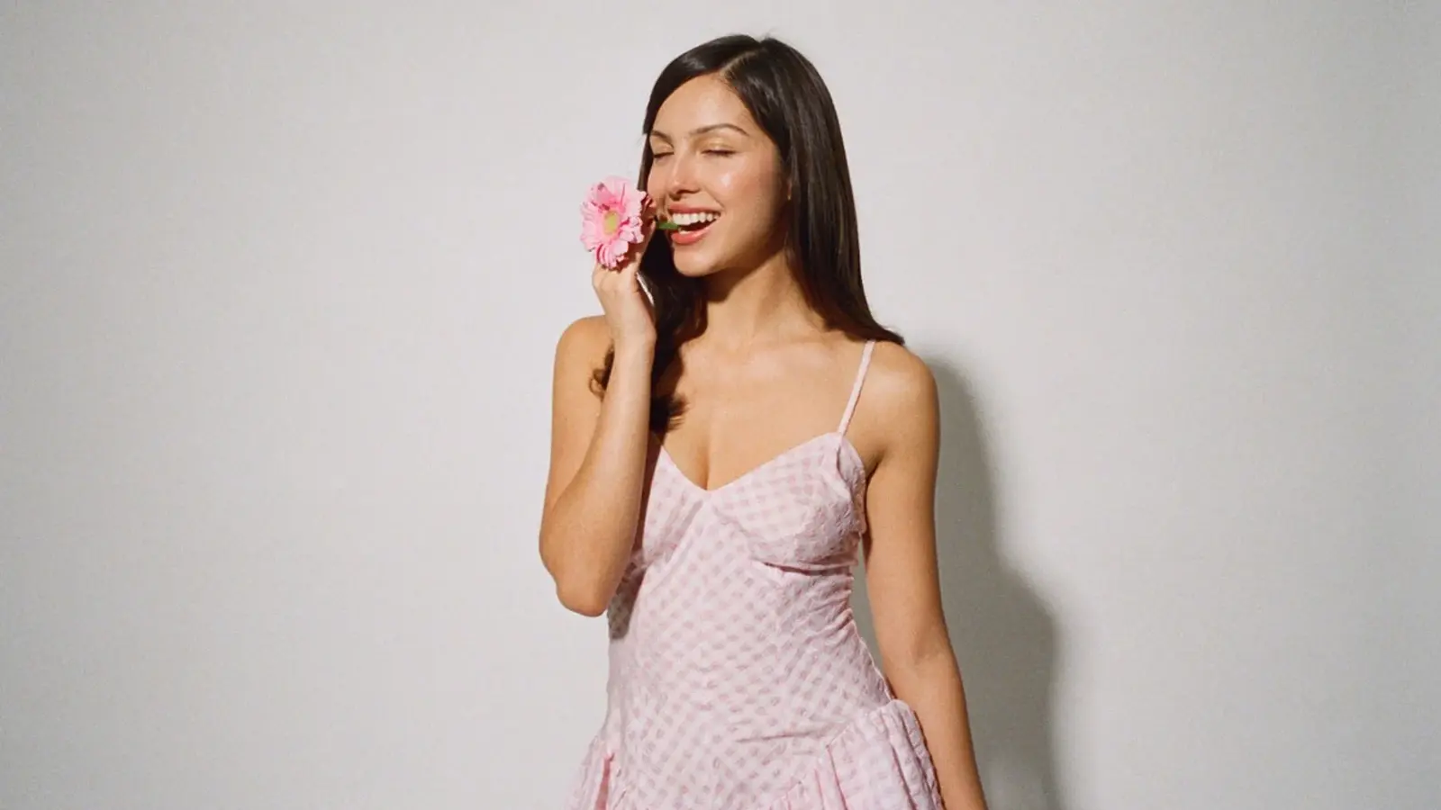Olivia Rodrigo in rosafarbenem Kleid hält eine Blume an ihr Gesicht vor neutralem Hintergrund, offizielles Pressefoto der Sängerin.