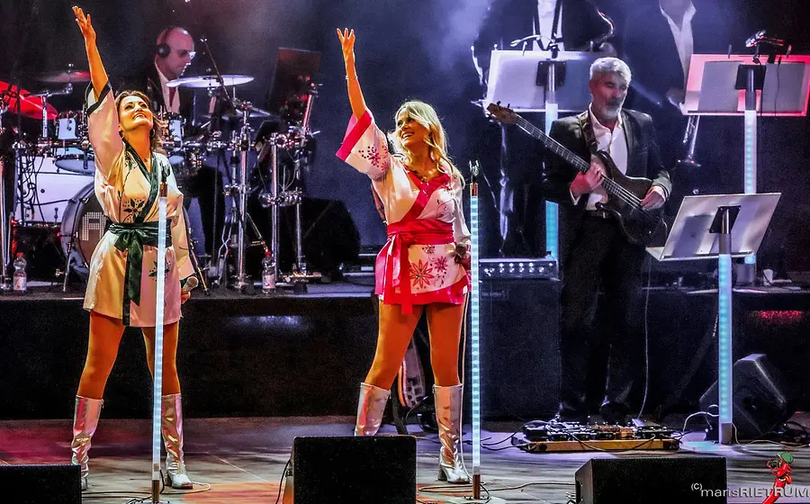 Bühnenfoto von One Night in Sweden mit zwei Sängerinnen in weißen Minikleidern mit roten und grünen Verzierungen im ABBA-Stil und silbernen Stiefeln vor Mikrofonständern, begleitet von Schlagzeug und Bass auf einer beleuchteten Konzertbühne.