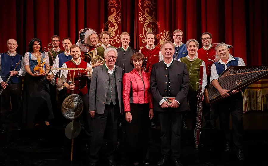 Gruppenfoto von Opern auf Bayrisch mit Ensemble aus Musikerinnen, Musikern und Mitwirkenden vor rotem Bühnenvorhang, zahlreiche Personen in Trachtenwesten mit Instrumenten wie Violine, Klarinette, Horn, Hackbrett und Schlagwerk auf einer Theaterbühne.