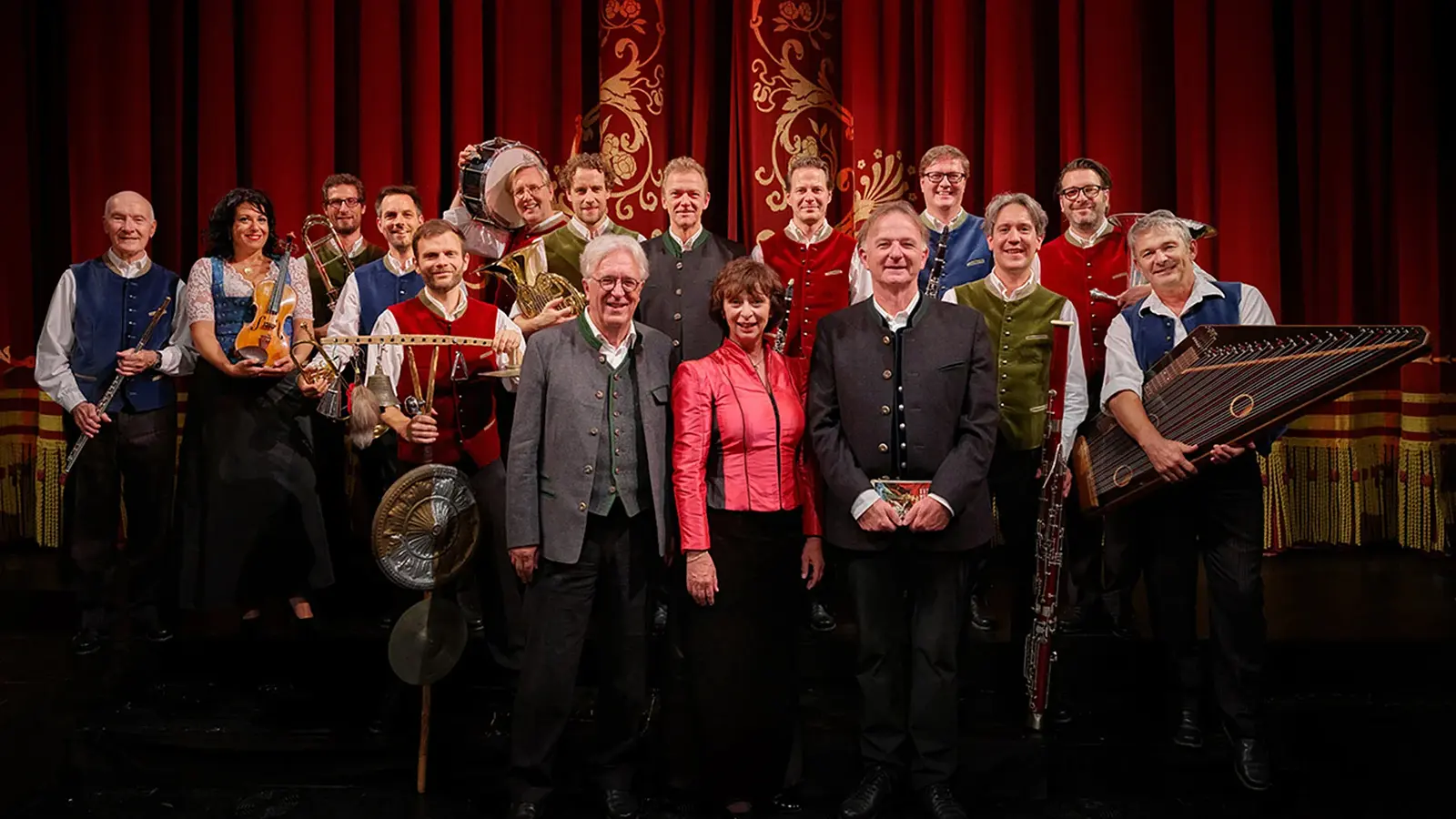 Gruppenfoto von Opern auf Bayrisch mit Ensemble aus Musikerinnen, Musikern und Mitwirkenden vor rotem Bühnenvorhang, zahlreiche Personen in Trachtenwesten mit Instrumenten wie Violine, Klarinette, Horn, Hackbrett und Schlagwerk auf einer Theaterbühne.