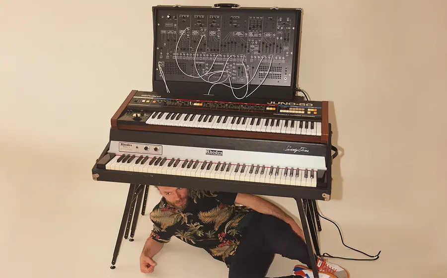 Porträtfoto mit Person in gemustertem Kurzarmhemd, die unter einem gestapelten Keyboard- und Synthesizeraufbau auf dem Boden sitzt, vor hellem Studiohintergrund mit Kabeln und elektronischen Instrumenten.