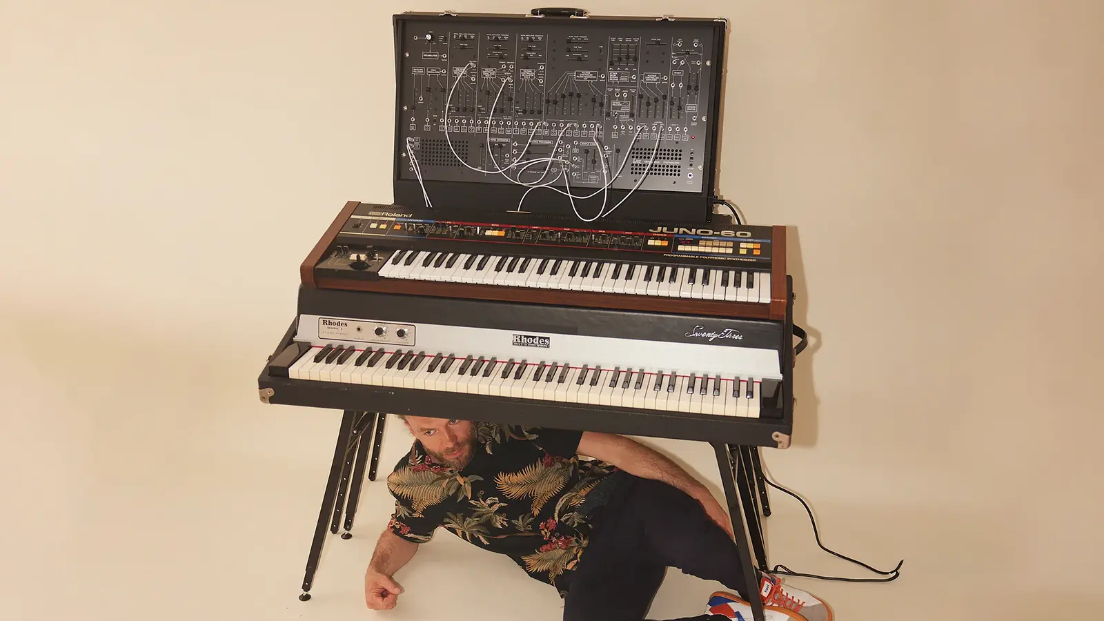 Porträtfoto mit Person in gemustertem Kurzarmhemd, die unter einem gestapelten Keyboard- und Synthesizeraufbau auf dem Boden sitzt, vor hellem Studiohintergrund mit Kabeln und elektronischen Instrumenten.