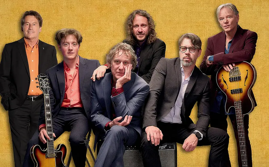 Sechs Mitglieder der Band Pepperland in dunklen Anzügen mit Instrumenten vor gelbem Hintergrund