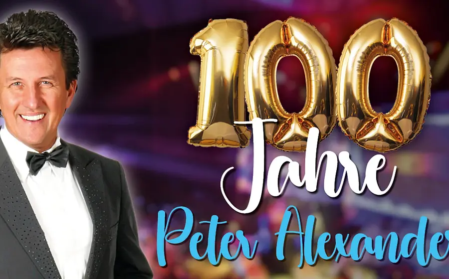 Peter Alexander im schwarzen Smoking vor Konzertkulisse mit goldenen „100 Jahre“-Ballons und Schriftzug zum Jubiläum.