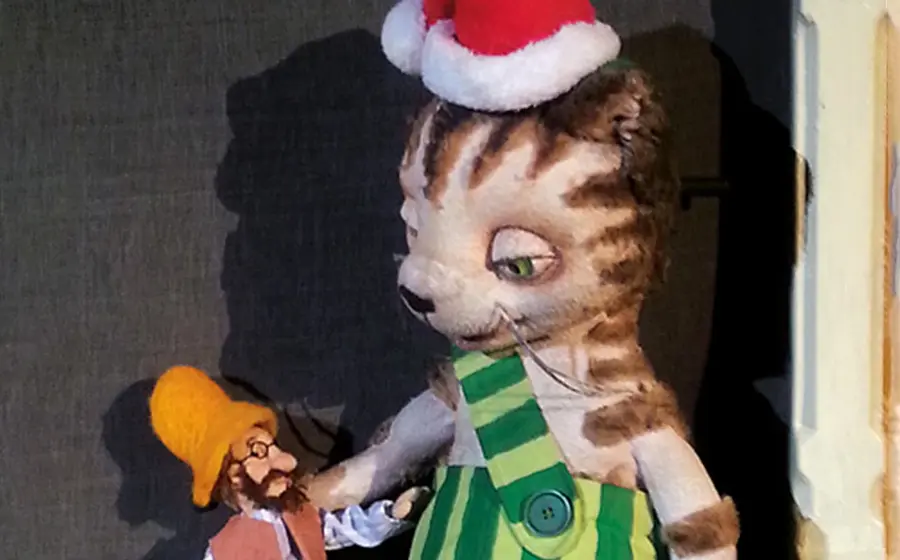 Bühnenfoto aus Pettersson kriegt Weihnachtsbesuch mit einer großen Katzenfigur in grün gestreifter Latzhose und Weihnachtsmütze, die einer kleinen Puppenfigur mit gelber Mütze gegenübersteht, vor dunklem Bühnenhintergrund.
