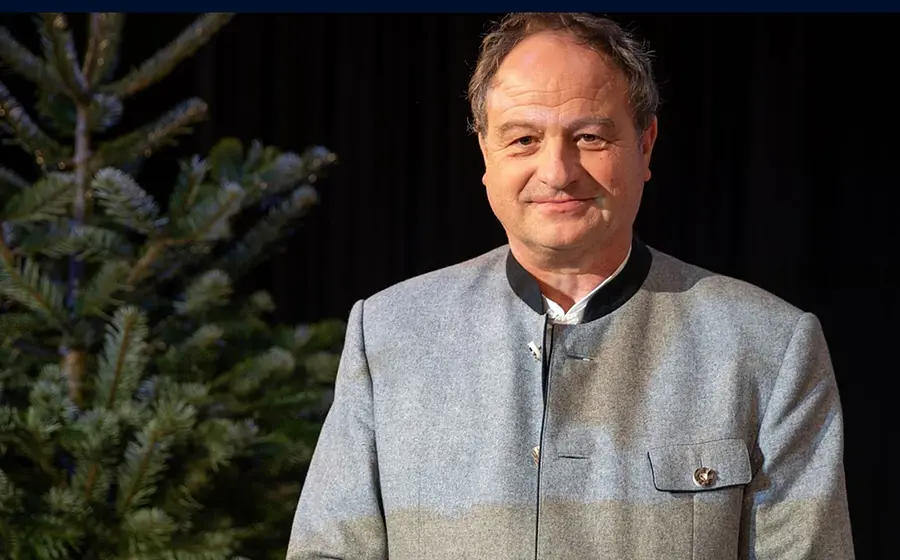 Rainer Maria Schiessler in grauer Trachtenjacke neben einem Tannenbaum vor dunklem Hintergrund