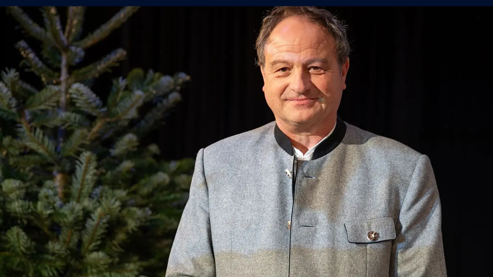 Rainer Maria Schiessler in grauer Trachtenjacke neben einem Tannenbaum vor dunklem Hintergrund