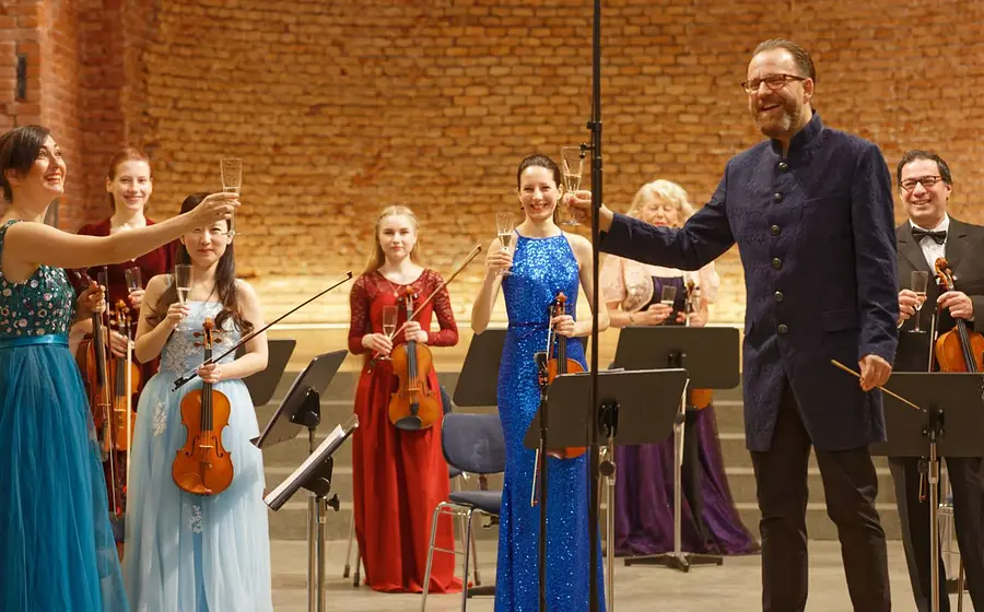 Bühnenfoto eines Kammerorchesters mit mehreren Musikerinnen und Musikern in festlicher Konzertkleidung, die Violinen und weitere Streichinstrumente halten, während eine Person im dunklen Gehrock und eine Person im blauen Abendkleid mit Sektgläsern anstoßen, vor Backsteinwand und Notenständern.