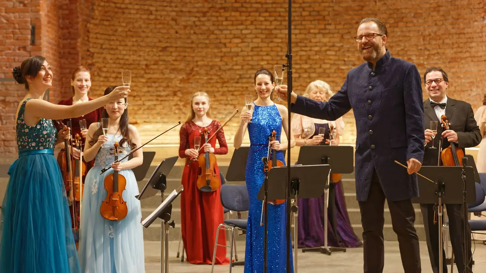 Bühnenfoto eines Kammerorchesters mit mehreren Musikerinnen und Musikern in festlicher Konzertkleidung, die Violinen und weitere Streichinstrumente halten, während eine Person im dunklen Gehrock und eine Person im blauen Abendkleid mit Sektgläsern anstoßen, vor Backsteinwand und Notenständern.