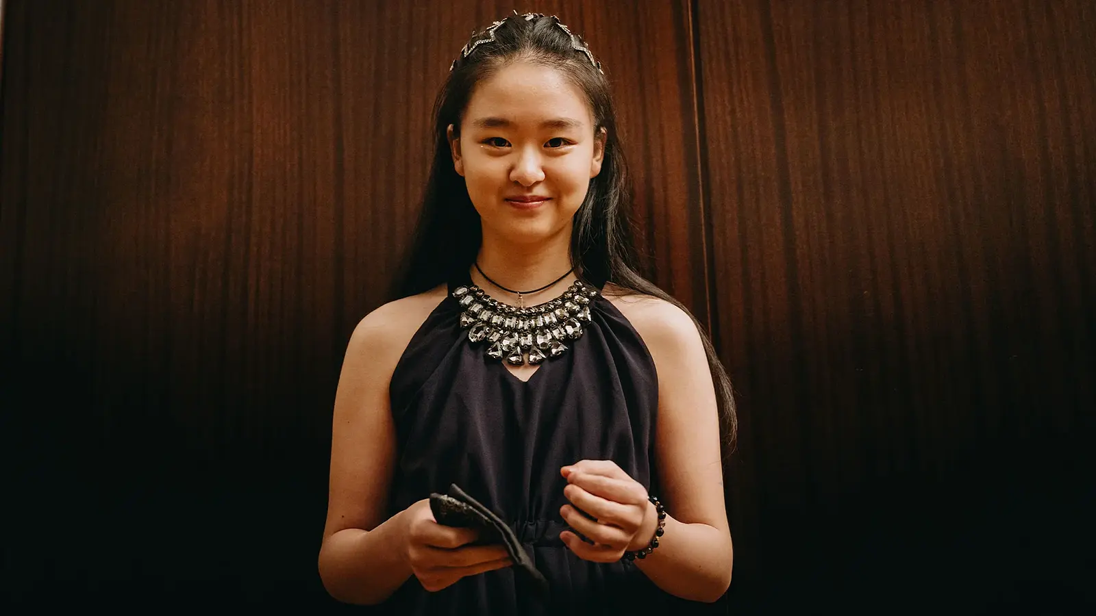 Die Pianistin Tianyao Lyu in schwarzer festlicher Kleidung mit dominanter Halskette lächelnd vor braunem Hintergrund