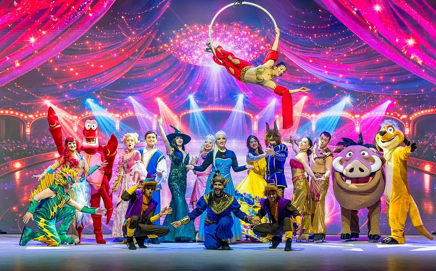 Bühnenfoto von Musical Dreams mit zahlreichen Darstellenden in farbenfrohen Kostümen bekannter Märchen- und Musicalfiguren, darunter eine Luftakrobatin am Ringtrapez, vor pink violett beleuchteter Showkulisse mit Lichteffekten