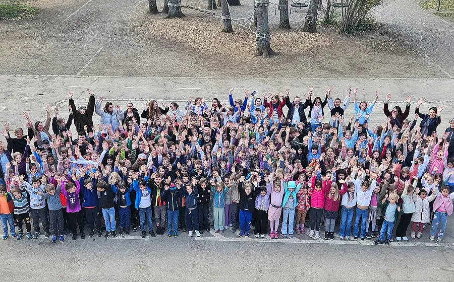 Große Gruppe von Kindern und Erwachsenen steht auf einem Schulhof und hebt die Arme in die Höhe. Im Hintergrund sind Bäume und ein Kletterbereich zu sehen