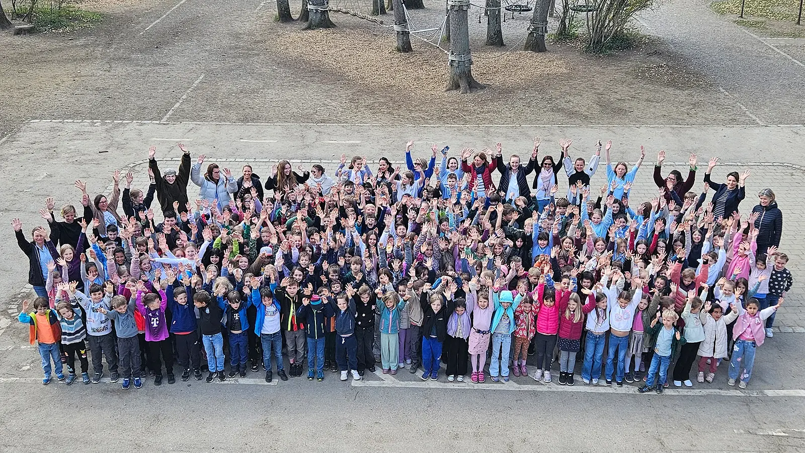 Große Gruppe von Kindern und Erwachsenen steht auf einem Schulhof und hebt die Arme in die Höhe. Im Hintergrund sind Bäume und ein Kletterbereich zu sehen