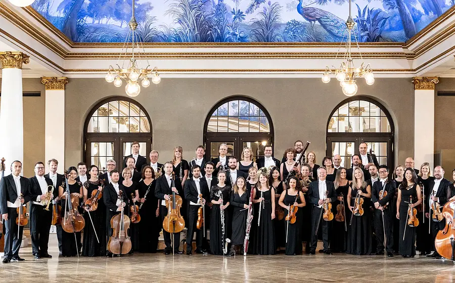 Großes Orchester in schwarzer Konzertkleidung steht mit Streich-, Blas- und Schlaginstrumenten in einem prachtvollen Saal mit hohen Fenstern, Säulen, Kronleuchtern und bemalter Decke.