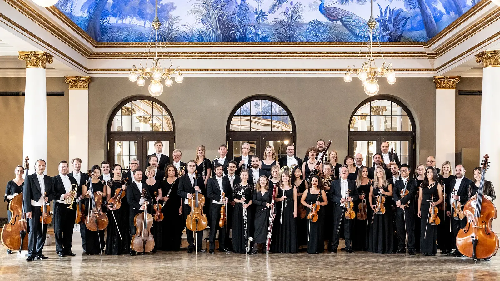 Großes Orchester in schwarzer Konzertkleidung steht mit Streich-, Blas- und Schlaginstrumenten in einem prachtvollen Saal mit hohen Fenstern, Säulen, Kronleuchtern und bemalter Decke.