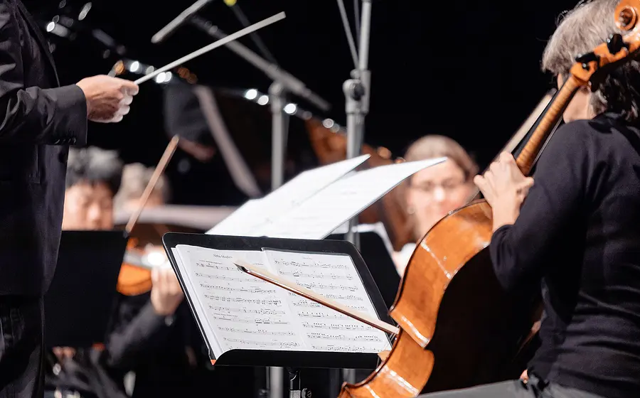 Nahaufnahme eines Orchesters während eines Konzerts. Im Vordergrund sind ein Violoncello, aufgeschlagene Notenblätter und ein Dirigierstab zu sehen, während weitere Musikerinnen und Musiker im Hintergrund spielen.