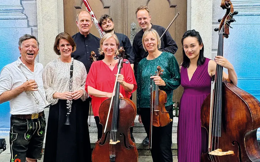 Gruppenfoto eines Ensembles mit sieben Personen, die vor einem Gebäude stehen und klassische Instrumente halten, im Hintergrund ein großes Holztor, links und rechts blau gefärbte Mauern