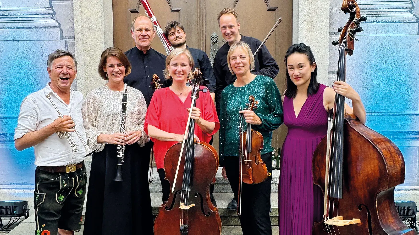 Gruppenfoto eines Ensembles mit sieben Personen, die vor einem Gebäude stehen und klassische Instrumente halten, im Hintergrund ein großes Holztor, links und rechts blau gefärbte Mauern