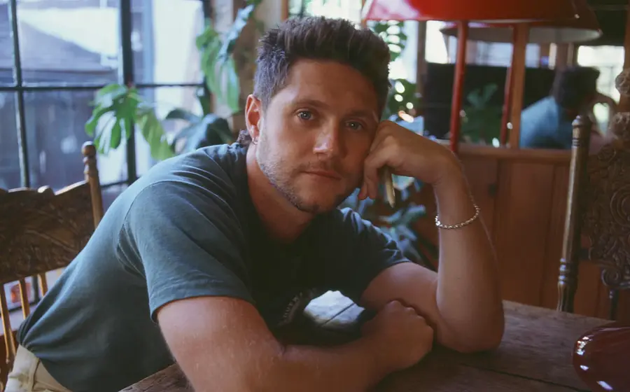 Niall Horan sitzt an einem Holztisch in einem lichtdurchfluteten Raum, stützt einen Arm auf den Tisch und den Kopf auf die Hand. Im Hintergrund befinden sich Holzmöbel, eine rote Lampe sowie mehrere Pflanzen vor einem großen Fenster.