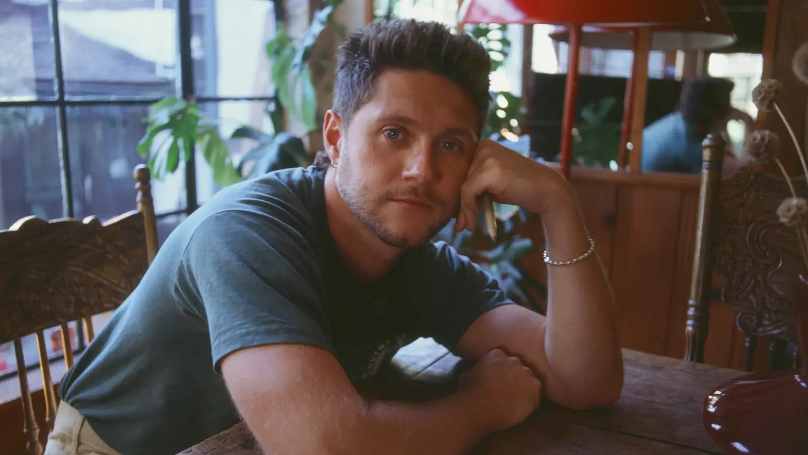 Niall Horan sitzt an einem Holztisch in einem lichtdurchfluteten Raum, stützt einen Arm auf den Tisch und den Kopf auf die Hand. Im Hintergrund befinden sich Holzmöbel, eine rote Lampe sowie mehrere Pflanzen vor einem großen Fenster.