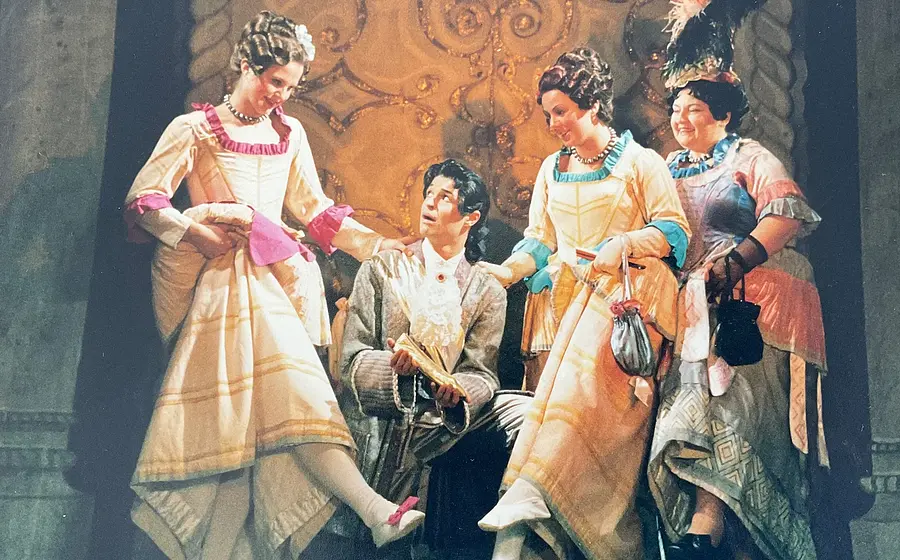 Historische Theaterszene mit vier Darstellenden in barocken Kostümen; drei Figuren in farbenfrohen Kleidern stehen um eine sitzende Person im silbernen Anzug, die ein Dokument hält, vor aufwendig verzierter Bühnenkulisse.
