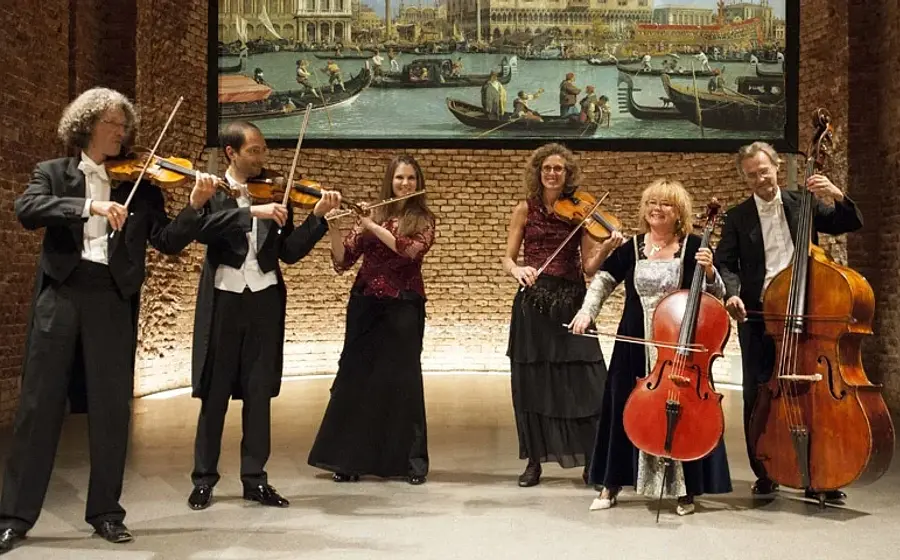 Pressefoto eines Kammerensembles mit fünf Musikerinnen und Musikern in Konzertkleidung vor einer Backsteinwand, mit Violinen, Viola, Violoncello und Kontrabass, darüber ein großformatiges Gemälde mit venezianischer Kanalszene