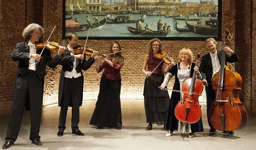 Pressefoto eines Kammerensembles mit fünf Musikerinnen und Musikern in Konzertkleidung vor einer Backsteinwand, mit Violinen, Viola, Violoncello und Kontrabass, darüber ein großformatiges Gemälde mit venezianischer Kanalszene