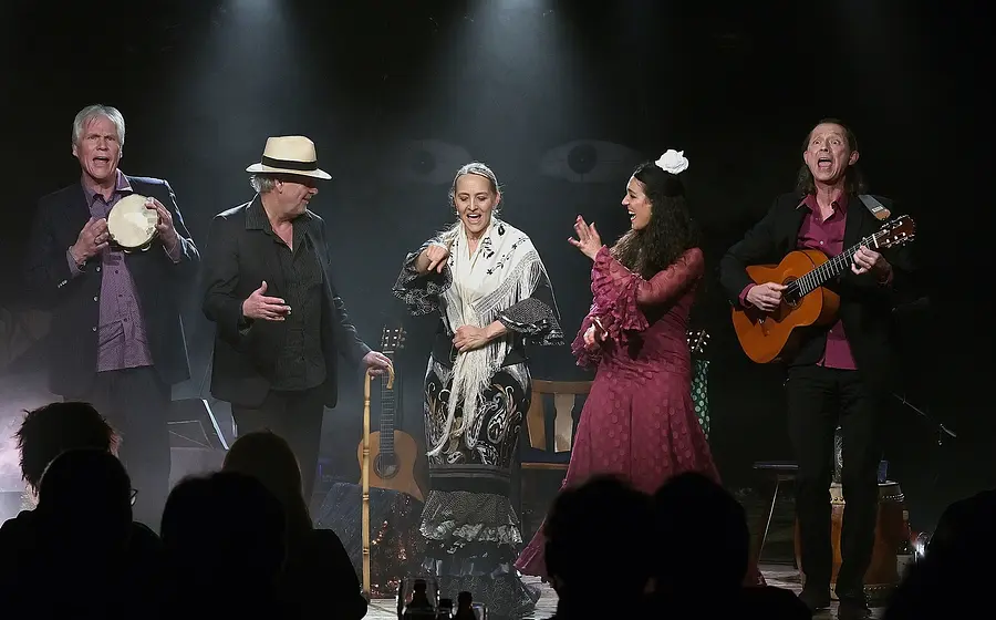 Fünf Musikerinnen und Musiker singen und musizieren auf einer Bühne mit Gitarre, Tamburin und Flamencoelementen vor Publikum.