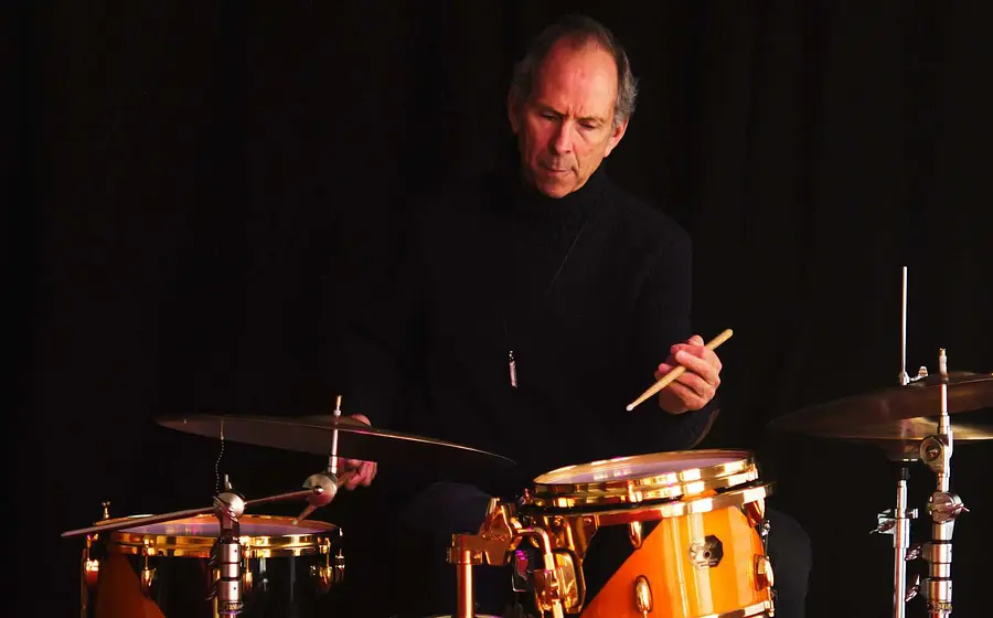 Rick Hollander in schwarzem Rollkragenpullover vor schwarzem Hintergrund, sitzend an einem Drumset mit Trommeln und Becken