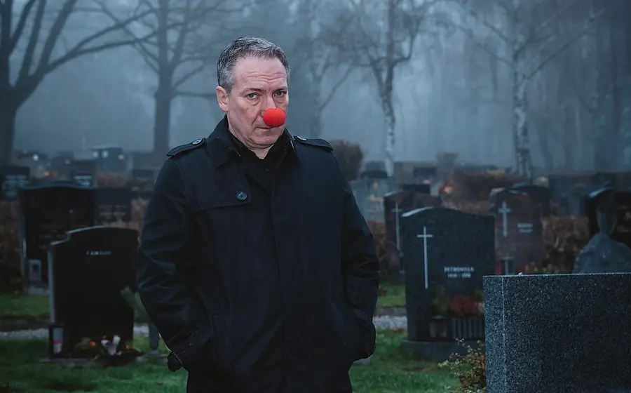 Der Künstler mit einer roten Clownsnase an einem Friedhof mit Grabsteinen