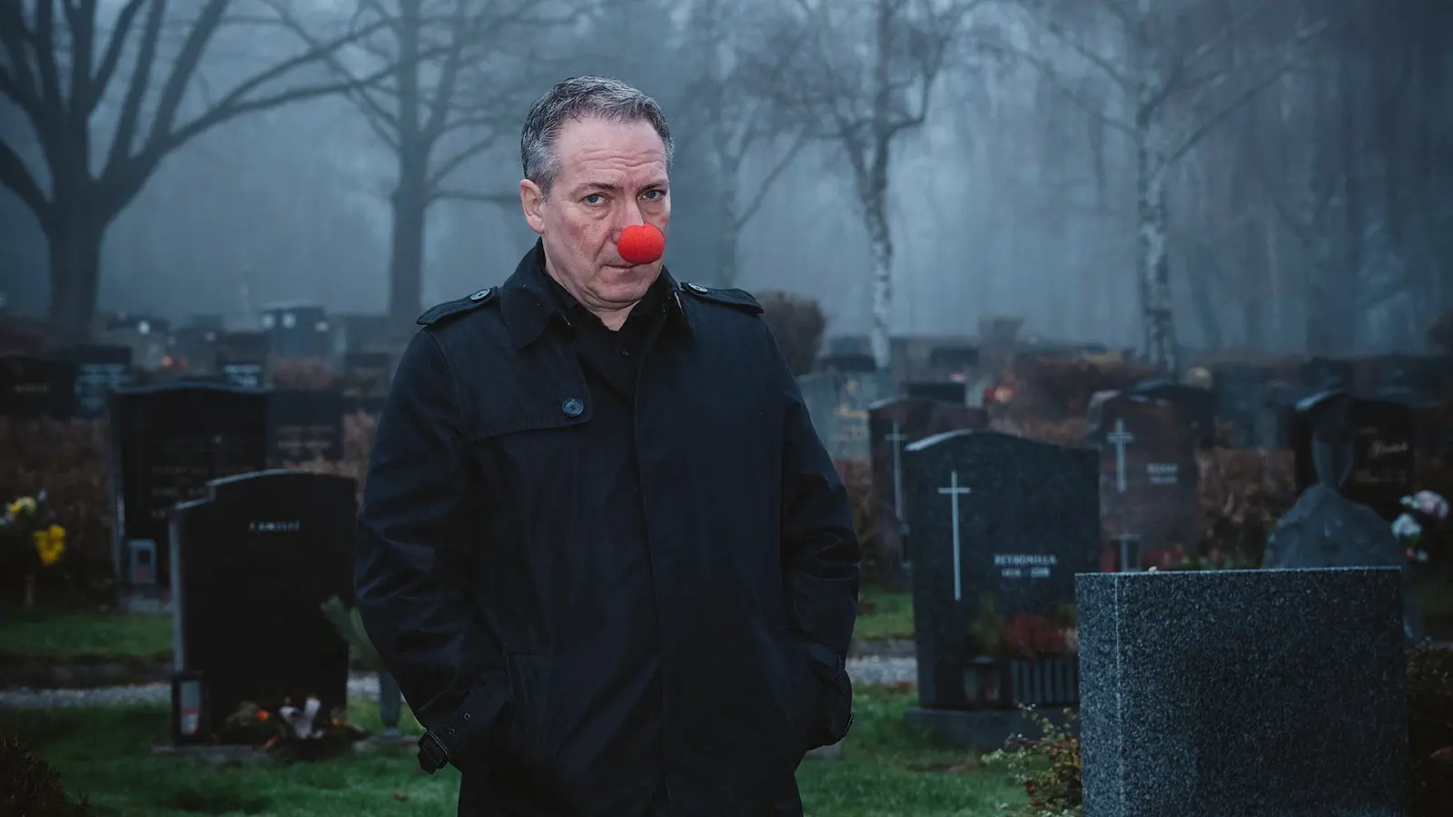 Der Künstler mit einer roten Clownsnase an einem Friedhof mit Grabsteinen